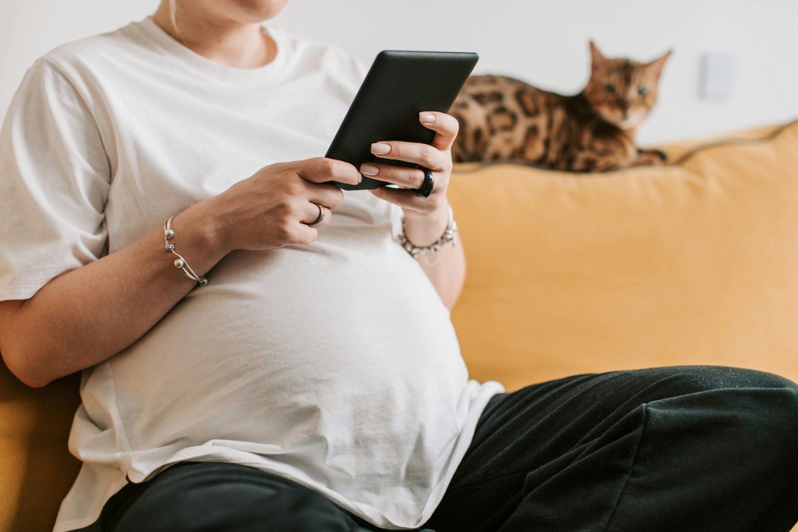 A pregnant woman using a tablet on a sofa with a Bengal cat beside her.